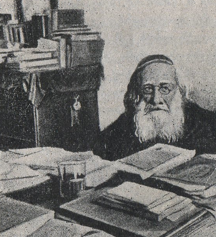 Photographie noir et blanc d'un homme agé portant des lunettes et une barbe blanche. Il est assis devant sa table de travail où se trouve de nombreux livres.