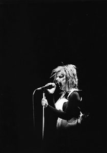 Photo noir et blanc d’une jeune femme, sur fond noir, devant un micro, bouche ouverte dans un cri, cheveux hirsutes