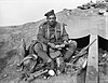 Black and white photograph of a man wearing military uniform sitting down. He is holding several bank notes and is surrounded by military equipment including a helmet, a rifle, several types of ammunition and soldiers' personal equipment.