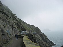 Photographie en couleurs d'un chemin taillé à flanc de falaise et délimité à droite par un parapet en pierre, un bâtiment installé à son extrémité.