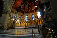 A view into the chancel is framed on the right by the lectern supported by a standing brass angel. The chancel is semi-circular and has a roof on wooden beams. The upper walls have brightly coloured mosaics of prophets and angels. The white marble altar and mosaic reredos of "The Last Supper" can be seen.