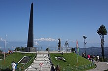 An obelisk on an elevated circular platform, with a few people standing around. Mountain peaks are visible in the background.