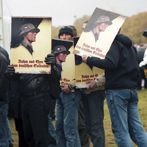 Color image of protesters in late 20th century clothing holding signs showing a soldier in Wehrmacht uniform