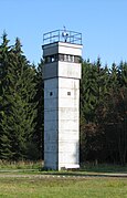 La torre cuadrada de color grisáceo en un prado, con árboles en el fondo. Solo la sección superior tiene ventanas; hay un reflector de techo plano de la torre, y la barandilla alrededor del borde exterior del techo.