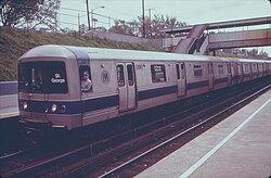 A Staten Island Railway train composed R44 subway cars on the Staten Island Railway. This image, taken in 1973, shows the cars with a since-removed blue stripe toward the bottom of the car body. The train is arriving at a platform to the left; the photo is taken from another platform to the right and in the foreground. The station is in a right-of-way below street level, and a covered footbridge connecting the two platforms is located to the right.