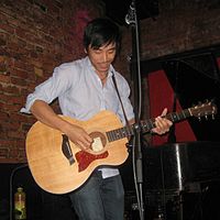 Alex Wong wearing blue jeans and open-necked button shirt, standing onstage in front of microphone, playing acoustic guitar