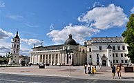 Seitliche Farbfotografie eines klassizistischen. weißen Gebäudes mit einem freistehenden Turm auf der linken Seite und einem viergeschossigen Gebäude mit verziertem Durchgang an der rechten Seite. Das mittlere Gebäude hat einen Säulengang und auf dem dunklen Dach sind zwei Eckkuppeln und zwei weiße Figuren mit einem Kreuz. Auf dem Platz im Vordergrund steht eine Figur mit einem Esel auf einem hohen Sockel.