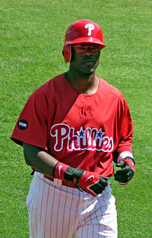 A baseball player wearing a red cap with the letter "P" inscribed. His jersey is also red, and is inscribed with the word "Phillies". His pants are white with thin red stripes running vertically across.