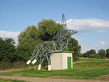 Skulptur, die einem Freileitungsmasten nachempfunden ist, der sich zum "Ausruhen" "hingesetzt" hat