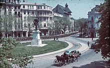 Period photo of thousands of armed soldiers marching down a city street and around a circular park, while a horse and carriage and car pass in the opposite direction