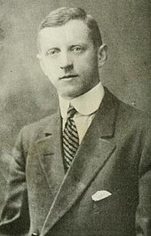 Black and white photo of a man wearing a dark suit jacket, a high-collared white dress shirt, and a striped necktie