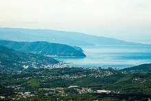 Vue couleur d'une zone urbaine, émergeant d'un espace vert boisé, au bord d'une étendue d'eau, sous un ciel blanc laiteux.