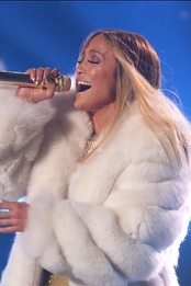 A brunette woman with a head set microphone, wearing a fur outfit, singing