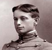 Head of a young man with neatly combed and parted hair wearing a military cadet's jacket, with cords running horizontally across the chest and a high, stiff collar.