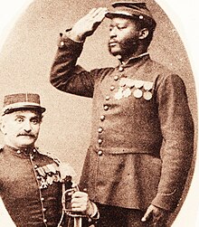 Photographie noir et blanc de deux soldats décorés en uniforme. Un, de couleur blanche, est assis et regarde de face. L'autre, de couleur noire, effectue le salut militaire.