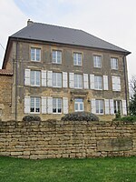 Photo en couleur d'une façade d’une imposante bâtisse en pierre comptant quatorze fenêtres, une porte d’entrée, une toiture grise ; le bâtiment est situé derrière une murette, également en pierre