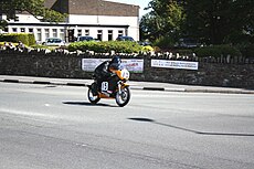 Ken Davis bij St Ninian's Crossroads tijdens de Junior Classic Manx Grand Prix in 2010. Op de achtergrond de laagbouw van St Andrews Church, achter de bomen rechts staat St Ninian's Church