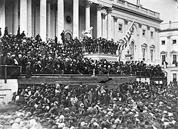 A large crowd in front of a large building with many pillars.