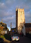 Square three stage stone tower. To the right is a building with a white wall and in the foreground a parked car.