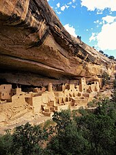 A color picture of a large sandstone ruins, with green trees below and blue sky above