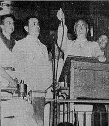 Black and white photo of a man (Roosevelt) behind a lectern holding the end of a wire above his head as others look on