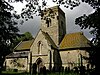 A stone church seen from the south, with a central tower in front of which is a two-storey gabled porch. To the left is a short nave, and to the right an apsidal chancel.