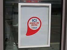 Photographie en couleurs d’une affiche blanche sur une vitrine. Au milieu de la feuille, le logo du réseau comporte l’inscription « Ici point de vente J’ybus ».