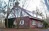 A house built of red stone house with a curved, sloping roof and white wooden top, seen from the side. It has brick chimneys at either end and some ivy on the visible side.