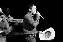 A man leaning on his left side, wearing a shirt and denim pants, holding a microphone in his left hand and a hat on the right.