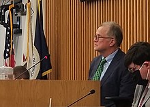 Dan Cronin seated on the dais of the board room. The flags of the United States, the State of Illinois, and County of DuPage are in the background.