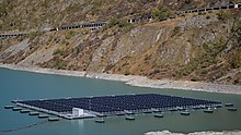 Au centre de la photo, on distingue des panneaux solaires flottant sur l'eau. En arrière plan, on aperçoit la paroi rocheuse plongeant dans le lac. On distingue une certaine hauteur de roche sans végétation indiquant que le lac de barrage n'est pas à son niveau le plus haut.