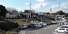 Blick auf einen niedrigen, modernen Gebäudekomplex, der leicht erhöht liegt und von Bäumen umstanden ist. Davor eine Betonfläche mit vielen parkenden Autos, in der Mitte der Fahrzeuge eine freie Fahrgasse mit weißen Pfeilen und einem Rollstuhl-Piktogramm.