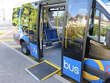 Photographie en couleurs d’un minibus bleu et noir en stationnement avec la rampe d’accès déployée et les portes ouvertes.