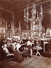 A black and white photograph of an elderly Victoria sat alongside a younger woman (Beatrice) reading a newspaper. The room is ornately decorated, with a number of photographs, paintings and a large chandelier hanging from the ceiling.