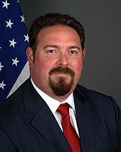 A photograph of a man with a goatee looking at the viewer while wearing a navy suit jacket, a white dress shirt, and a red necktie