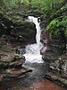 A plume of water falls in a narrow channel carved in layers of reddish brown rock. The falls, surrounded by green foliage, spill into a shallow pool.