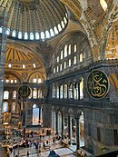 A photograph taken from a height of the interior of a domed building built in the classical style, large circular shield like objects with Arabic calligraphy on them are on the walls