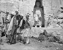 Black and white photograph of 6 Afghan men. Behind them, a giant sculpture is carved into the mountain side.