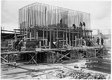 Scaffolding surrounds a half-finished concrete foundation. Dozens of metal steel poles rise from the foundation. A dozen workmen are visible and involved in various construction tasks.