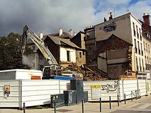 Un engin de chantier, derrière des palissades métalliques, procède à la destruction d'un bâtiment.