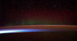 Earth's night-side upper atmosphere appearing from the bottom as bands of afterglow illuminating the troposphere in orange with silhouettes of clouds, and the stratosphere in white and blue. Next the mesosphere (pink area) extends to the orange and faintly green line of the lowest airglow, at about one hundred kilometers at the edge of space and the lower edge of the thermosphere (invisible). Continuing with green and red bands of aurorae stretching over several hundred kilometers.