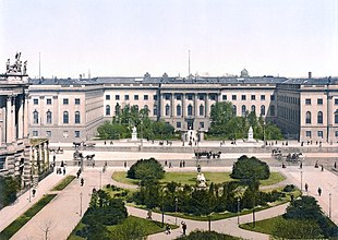 Frontale Farbfotografie in der Obersicht eines dreigeschossigen Gebäudes im Rokoko-Stil mit zwei Seitenflügeln. Der Mittelteil ist mit Säulen verziert und vor dem Zaun stehen zwei Marmorfiguren auf Sockeln. Auf der breiten Straße, die in der Bildmitte waagerecht verläuft, fahren Kutschen. Im Vordergrund ist ein großer Platz mit Rasen und Bäumen zu sehen.