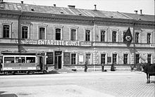 Frontale Schwarzweißfotografie von einer zweigeschossigen Häuserfassade mit dem Banner der Ausstellung „Entartete Kunst“. Links fährt eine Straßenbahn und rechts stehen einige Menschen. Am Eingang befinden sich weitere Tafeln und eine Hakenkreuzflagge hängt am oberen, rechten Fenster.