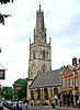 A stone church in a town seen from the southeast. St the far end is a large tower surmounted by a truncated spire with pinnacles and, at the top, a ball finial