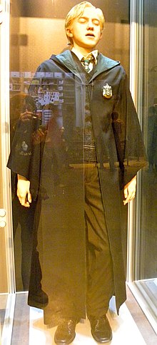 Farbfotografie einer Figur in einer Vitrine, die die Augen geschlossen hat. Sie ist ein Junge und trägt blondes Haar und einen langen schwarzen Umhang mit einem Wappen als Aufnäher.