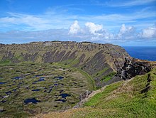 Colour photo from a cliff top down onto a pond strewn marsh backed by high cliffs, an area of sea is visible through a gap in the cliffs.