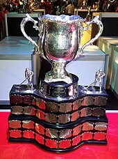 Silver bowl trophy with two large handles, mounted on a wide black plinth engraved with team names on silver plates