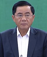 Trần Cẩm Tú standing in front of a green background, wearing glasses, a black suit and a white shirt.