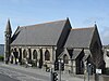 A steep-roofed, grey stone Gothic-style church with a prominent turret at the far corner, under sunny skies