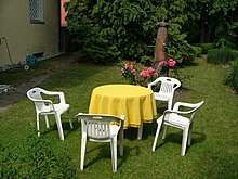 In einem privaten Garten auf grüner Wiese steht eine sitzgarnitur aus vier weißen Plastikstühlen und einem runden Tisch mit gelber Decke